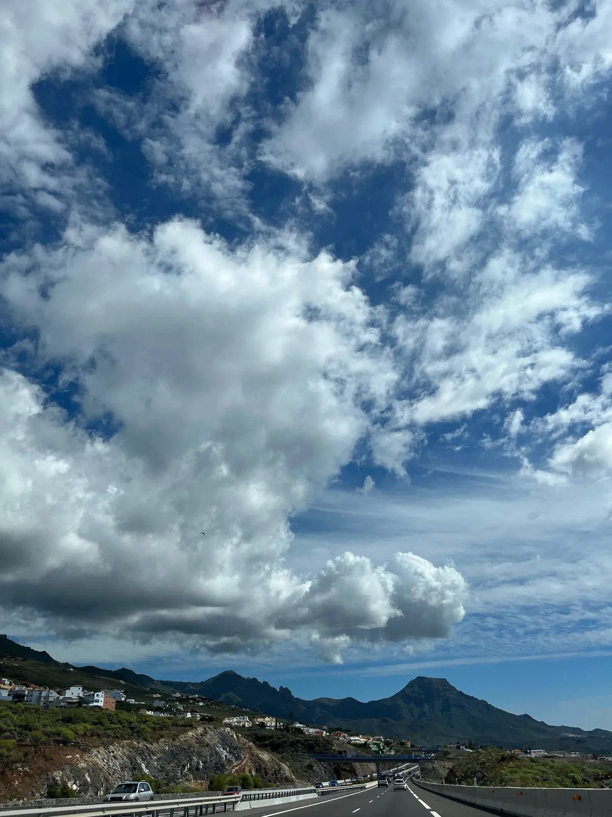 Highway with cars beneath towering clouds, with mountains and hillside houses