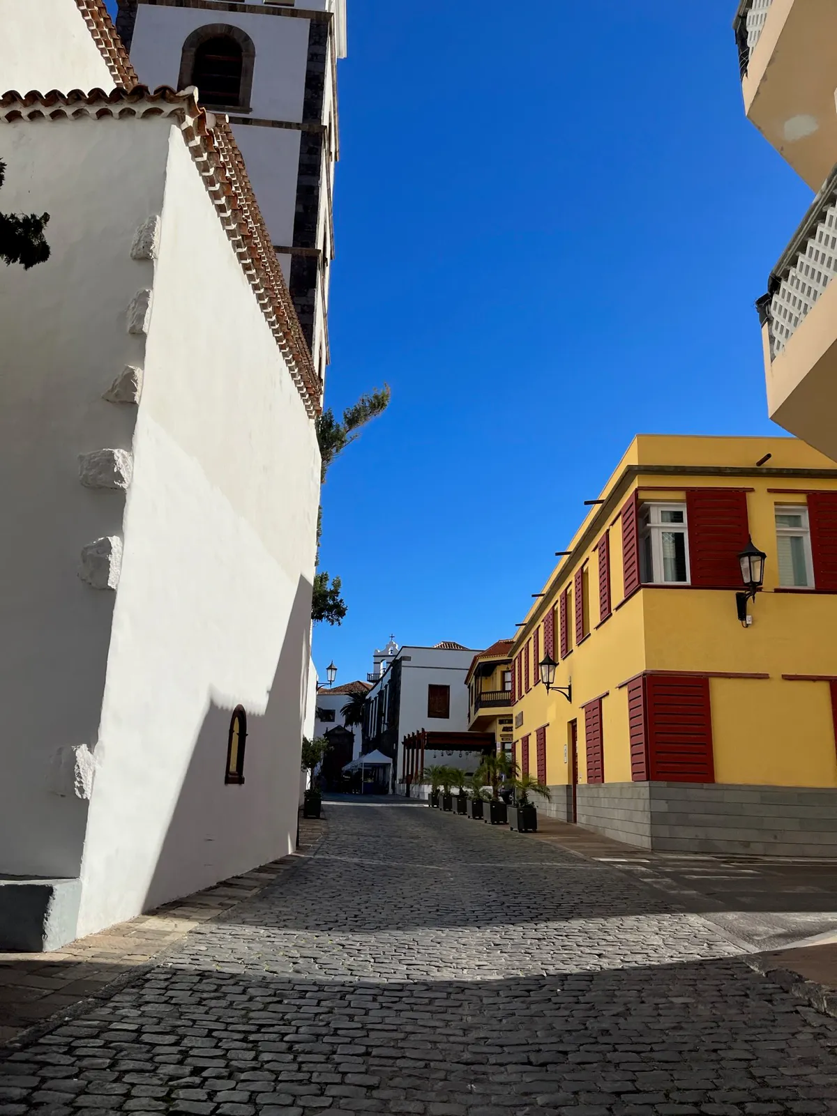 Cobbled lane beside a white church tower and a yellow house with red shutters.