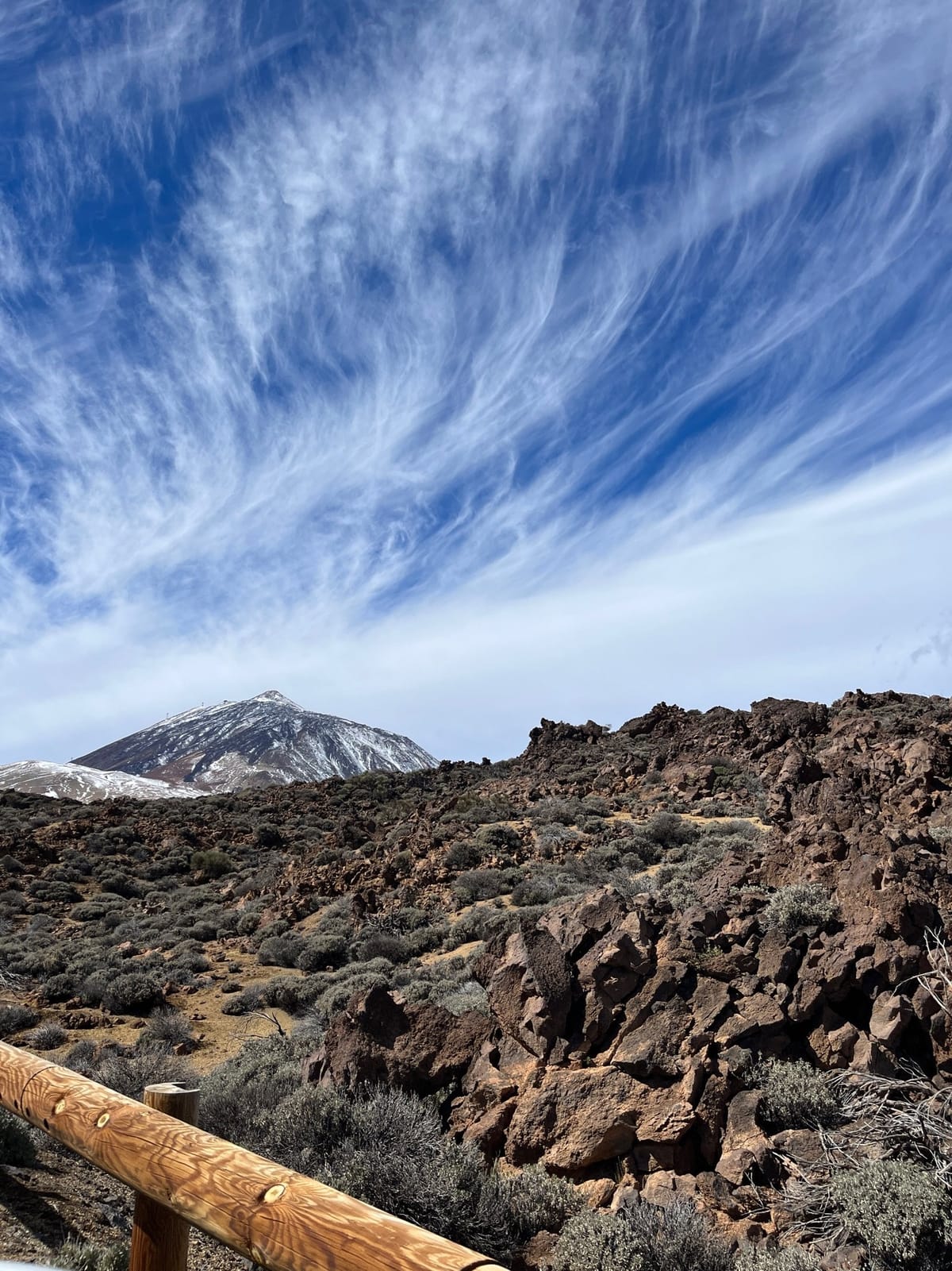 Snow-capped Teide beyond dark lava rocks, with a wooden rail in front