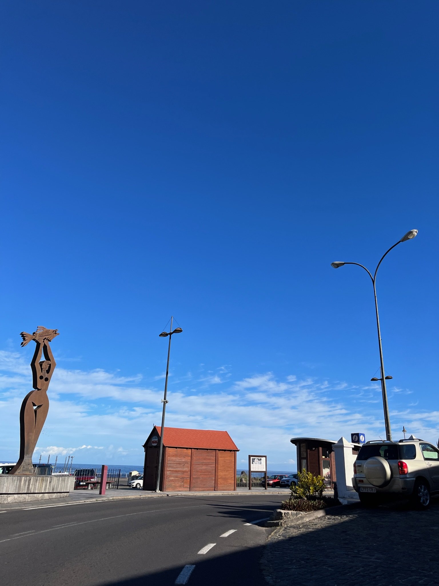 A metal sculpture of a figure lifting a fish beside a waterfront road.