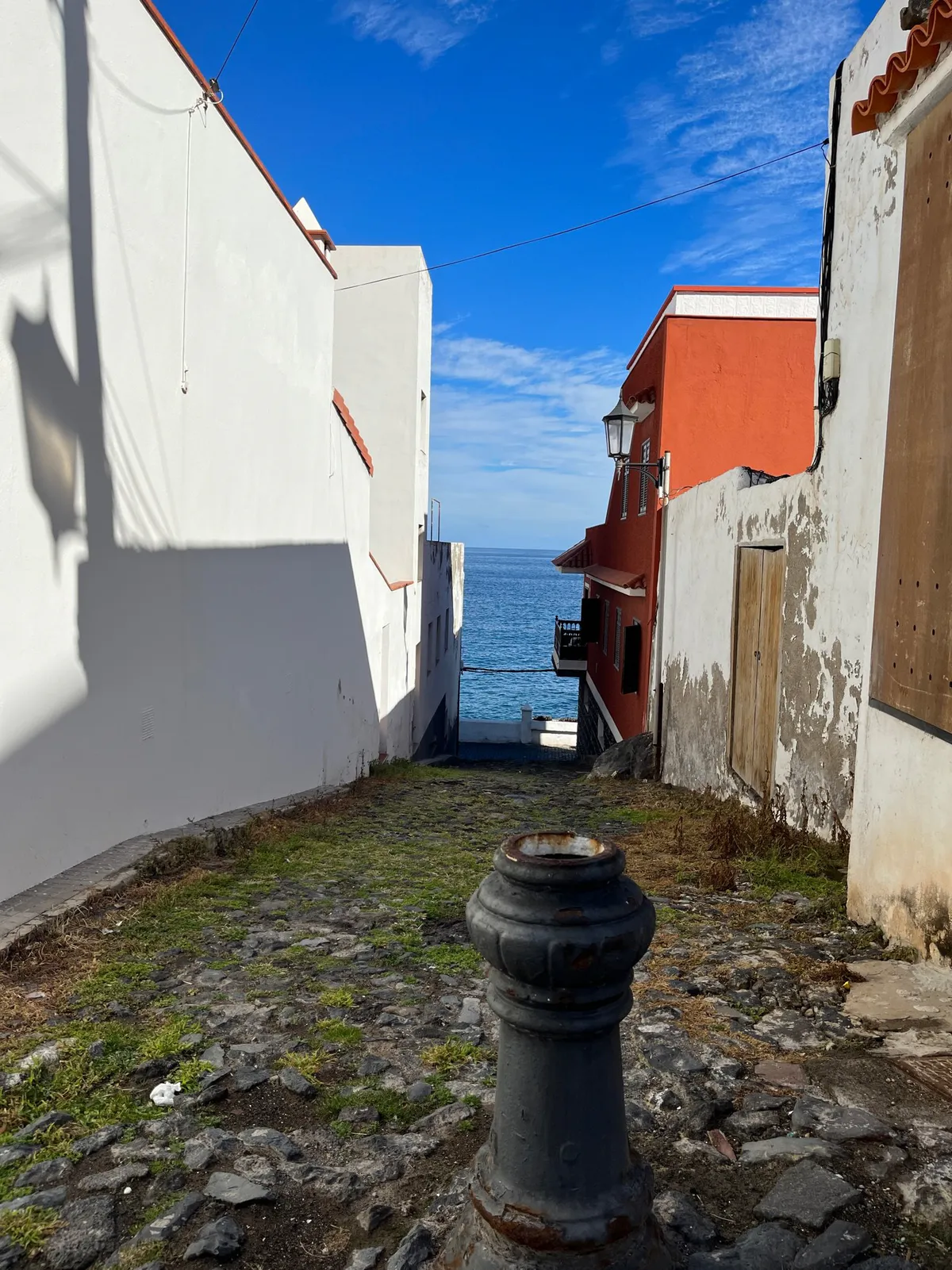 A metal post in a steep alley between white and orange houses, opening to the sea.