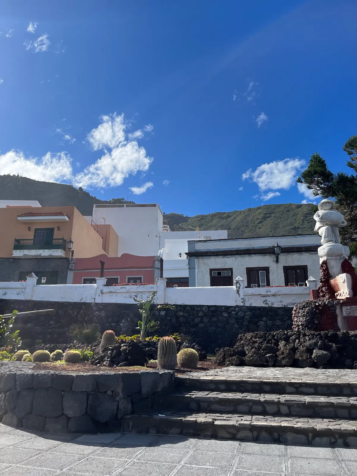 A cactus bed and stone steps below pastel houses, with a white statue at the right.