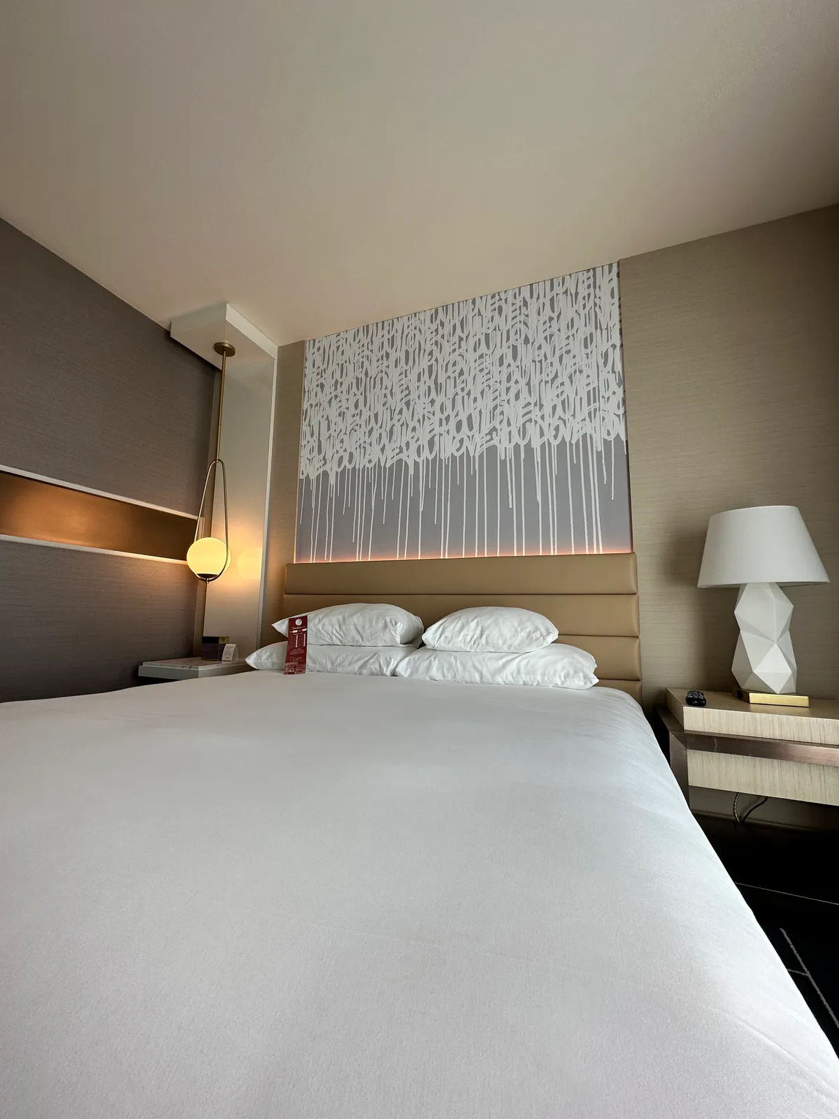 View across the white duvet toward pillows, a pendant, and a bedside lamp