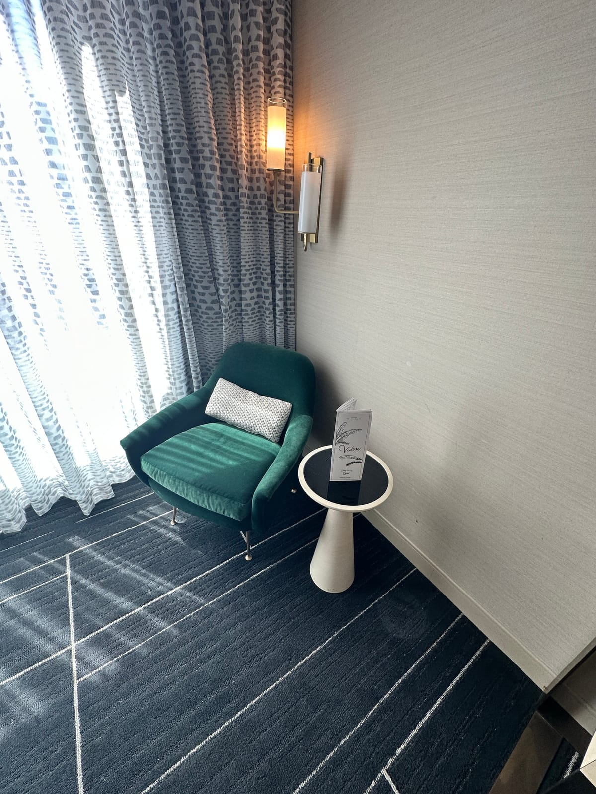 Green velvet chair and small table by patterned curtains and a wall sconce