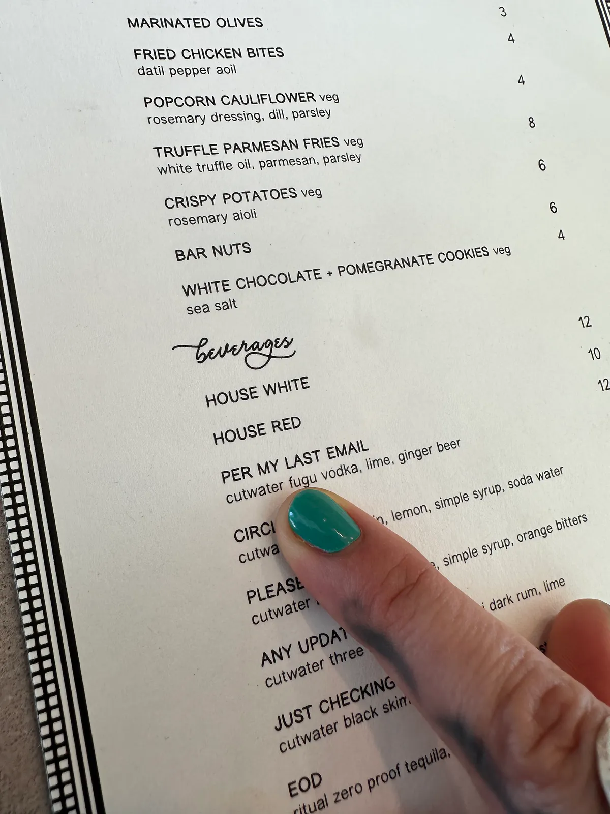 Teal-painted fingernail pointing at a cocktail name on the drink menu