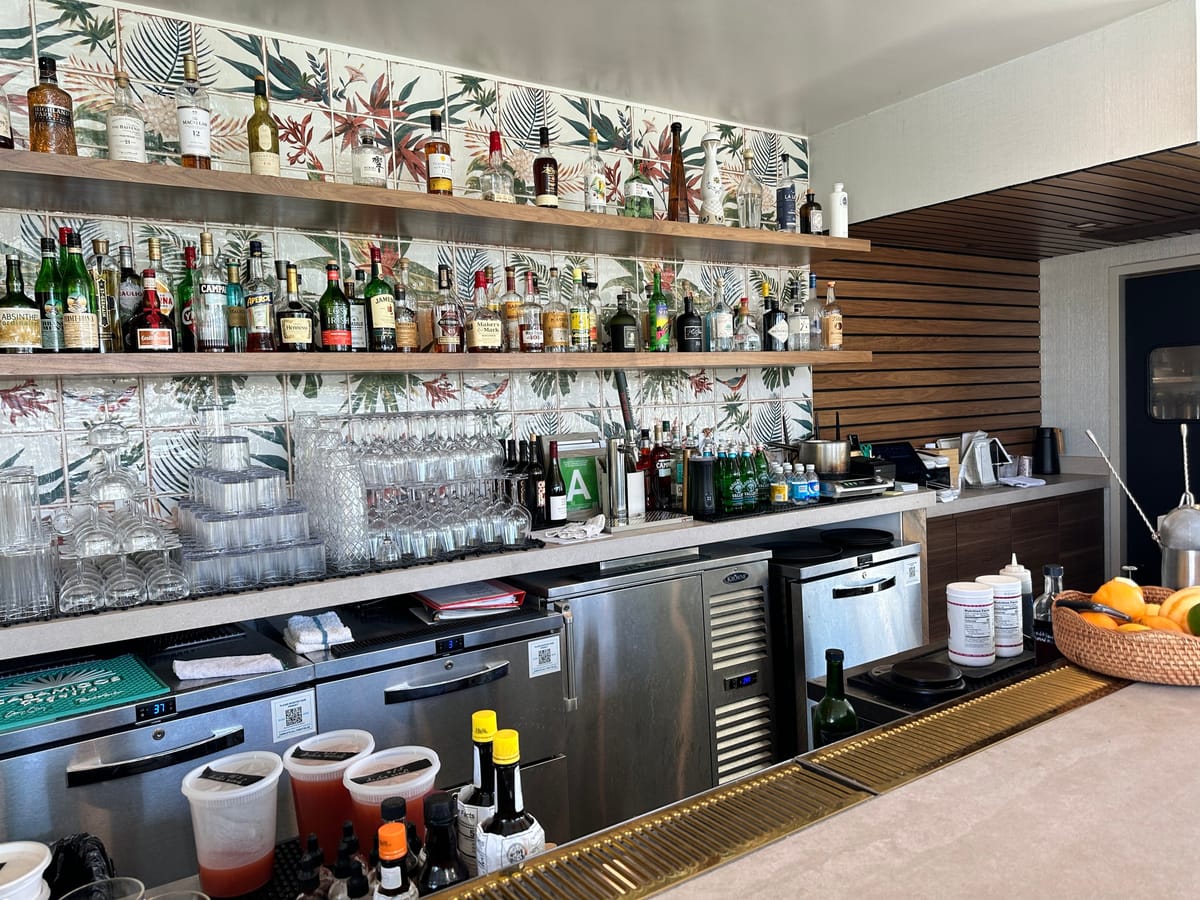 Bar shelves packed with liquor bottles and stacked glasses against tropical leaf tile