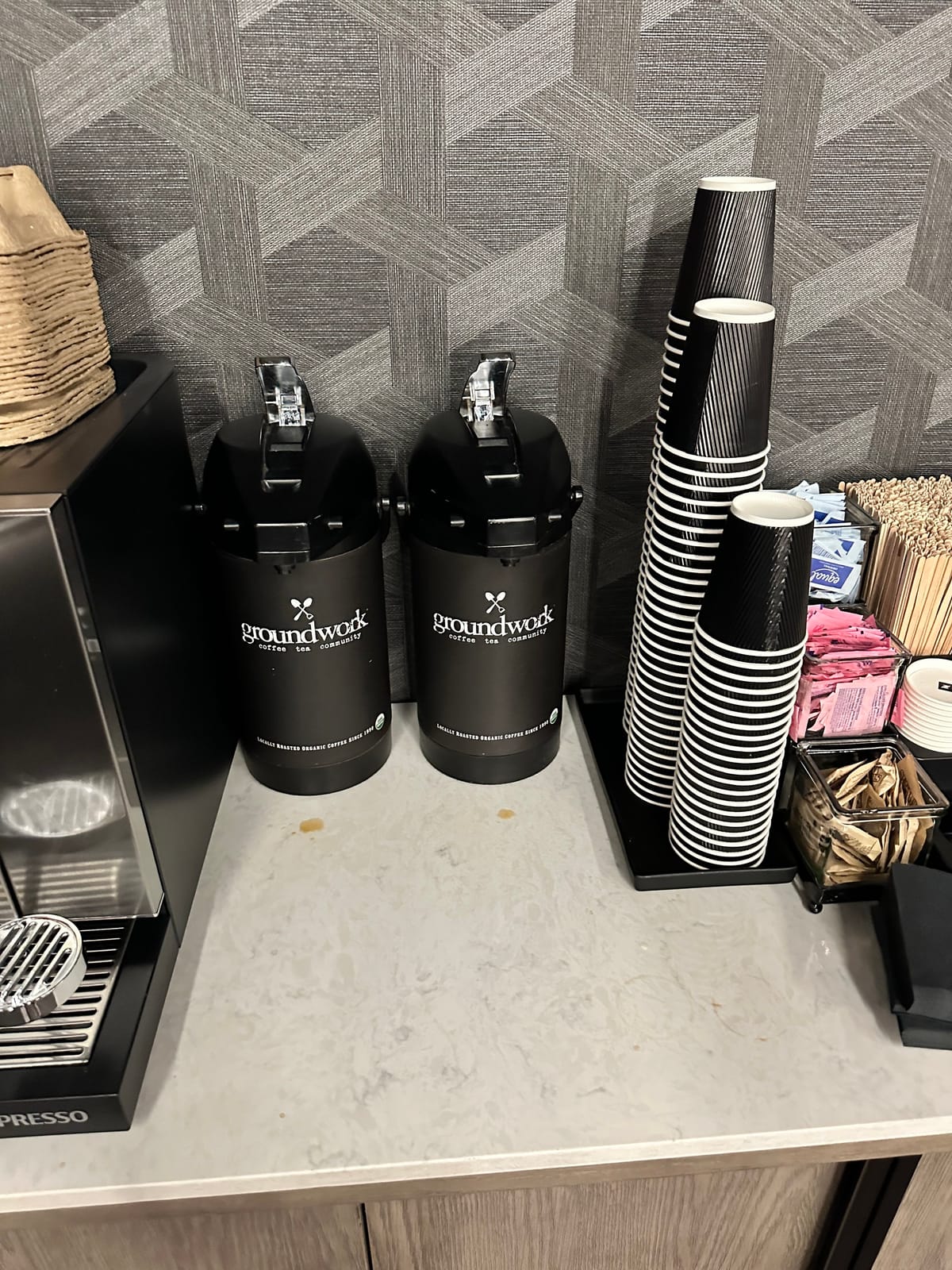 Two black Groundwork airpots beside tall stacks of striped paper cups