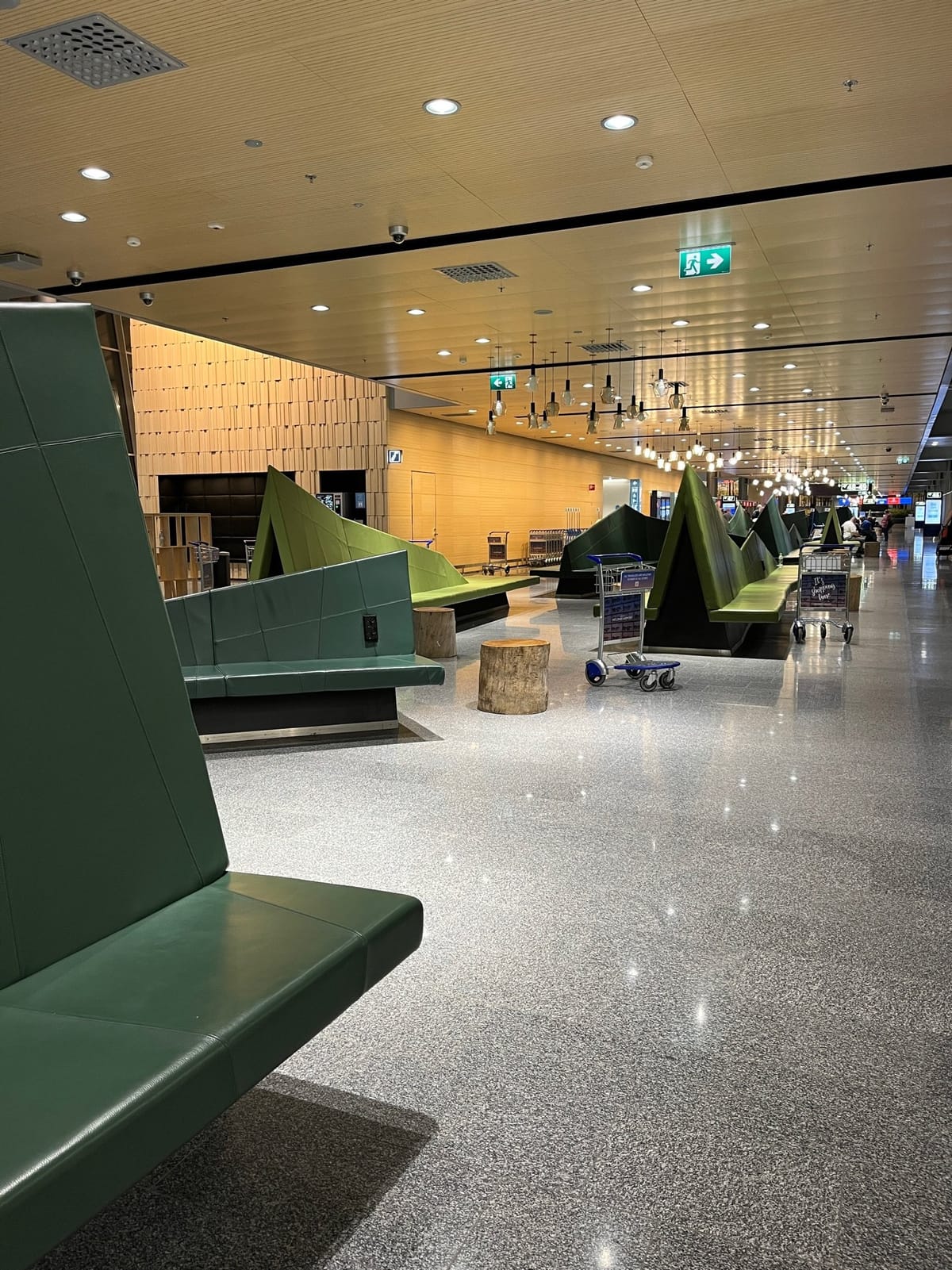 A corridor of angular green seating, log stools, and parked luggage carts