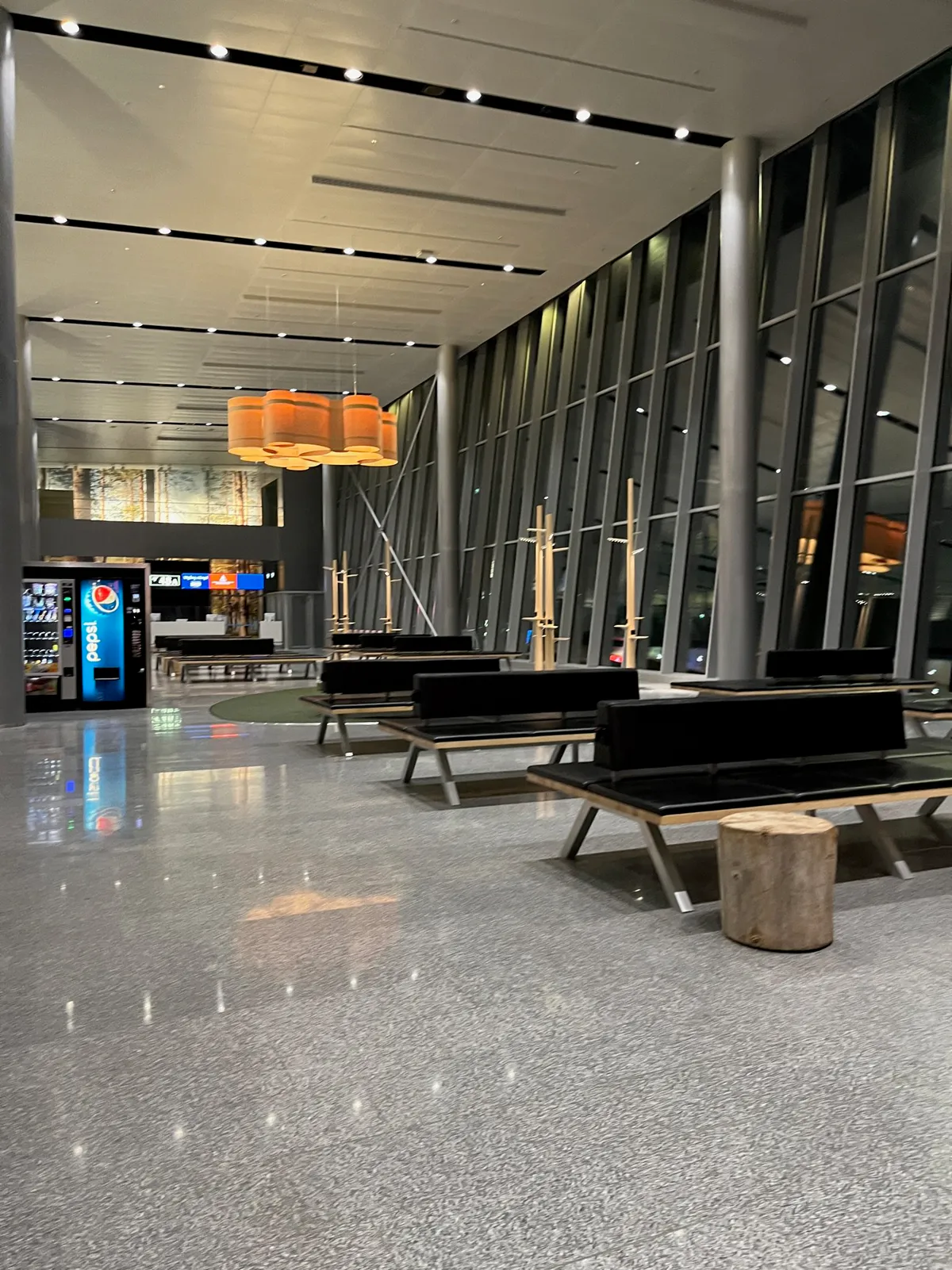 Black benches and log stools under orange barrel lights beside tall terminal windows