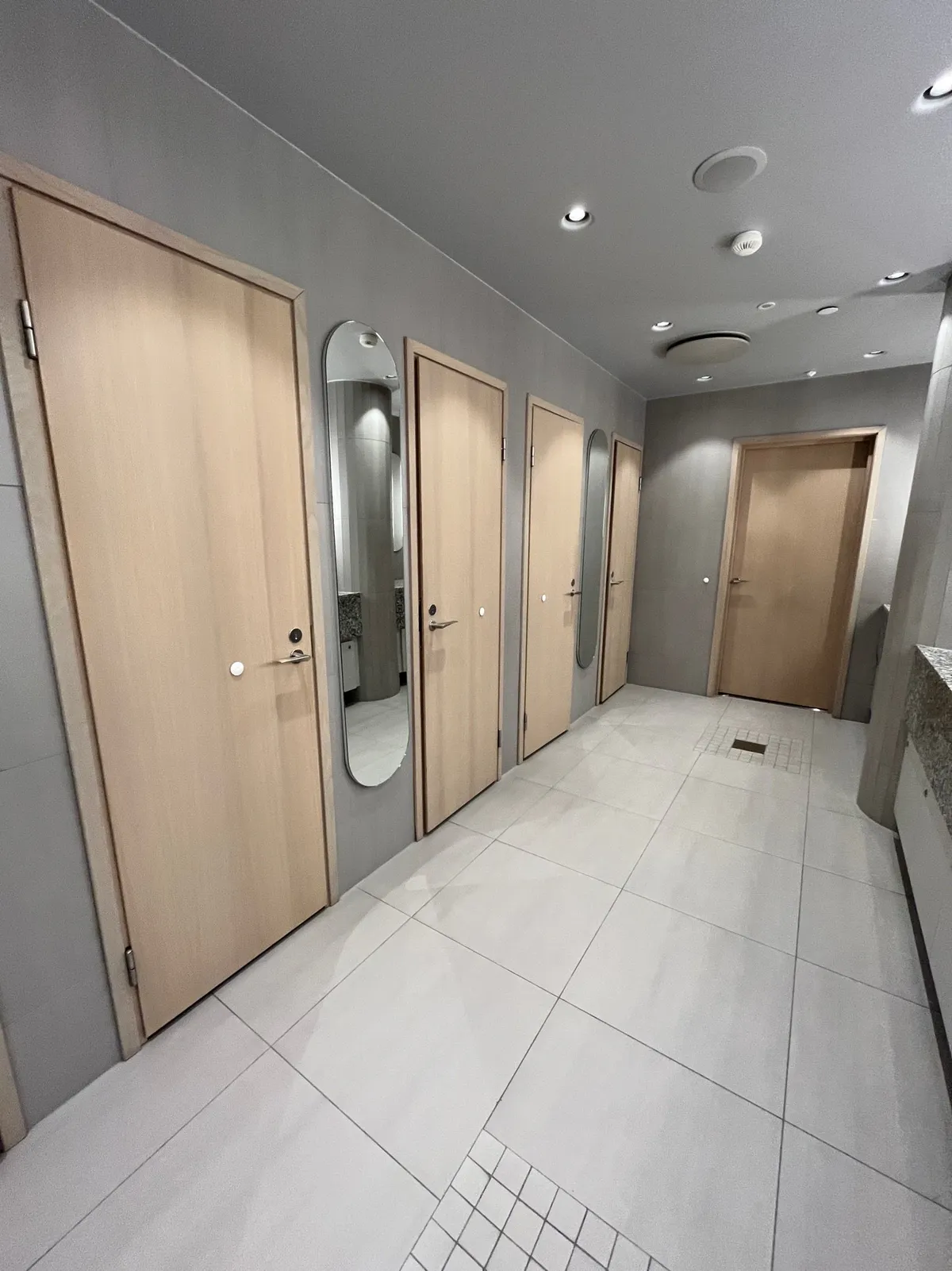 A restroom hallway lined with light wood stall doors and oval mirrors