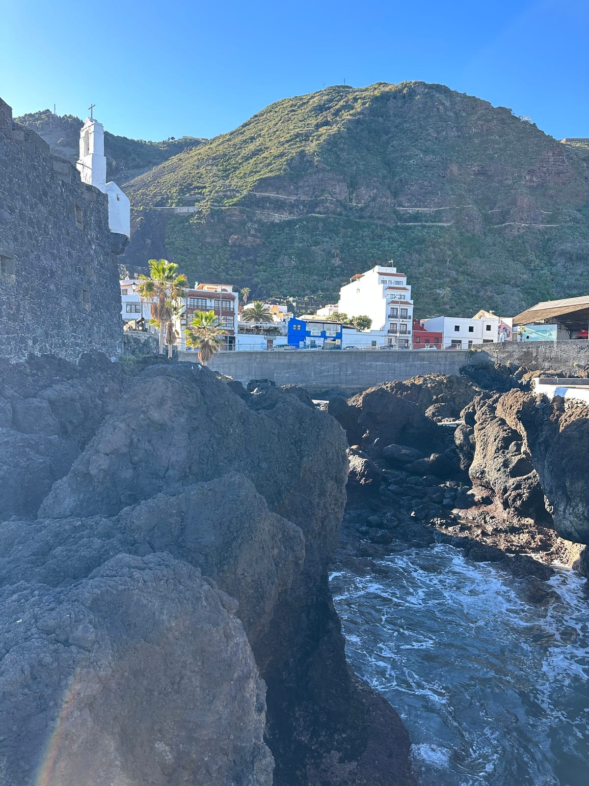 Dark volcanic rocks and surf below a stone tower, waterfront houses, and a green mountain.