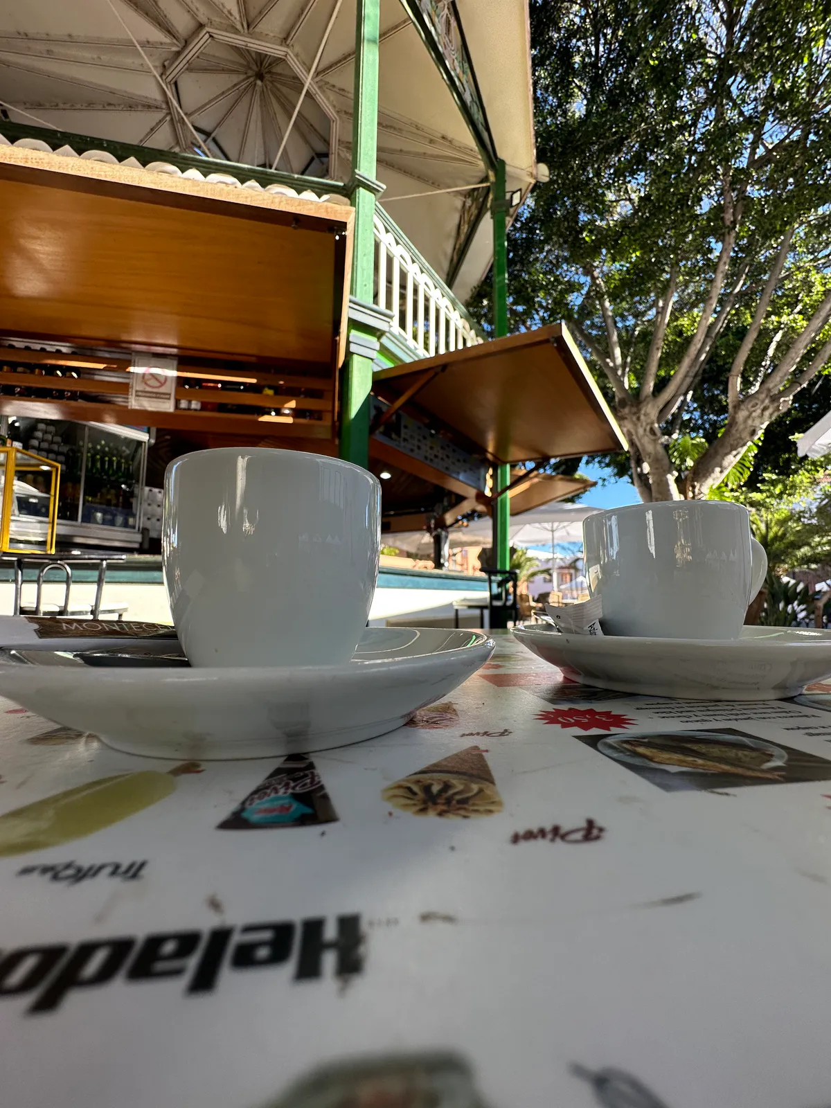 Two white coffee cups on a menu table at a shaded outdoor cafe terrace.