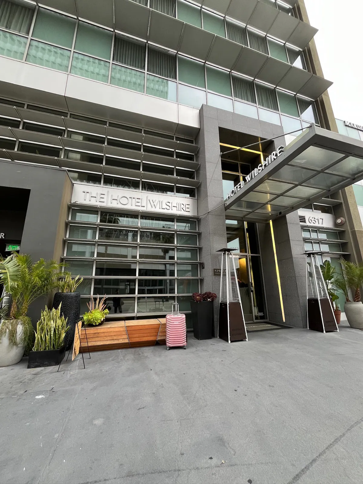 Front hotel entrance with a pink suitcase, planters, and a wood bench