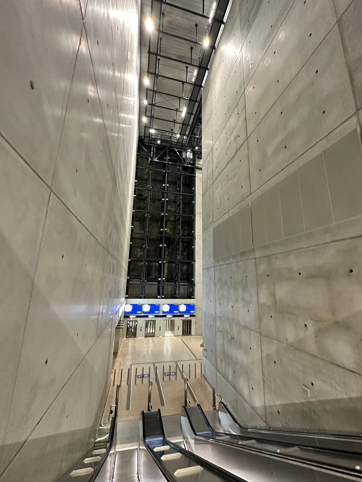 Escalators descending through a tall concrete atrium toward a row of blue screens