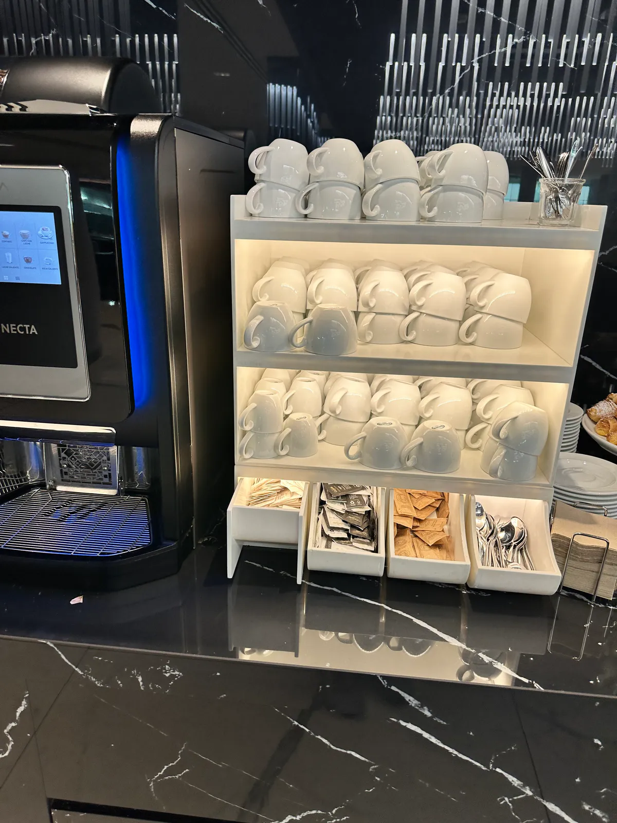 Lounge coffee machine beside stacked white cups, tea packets, and spoons