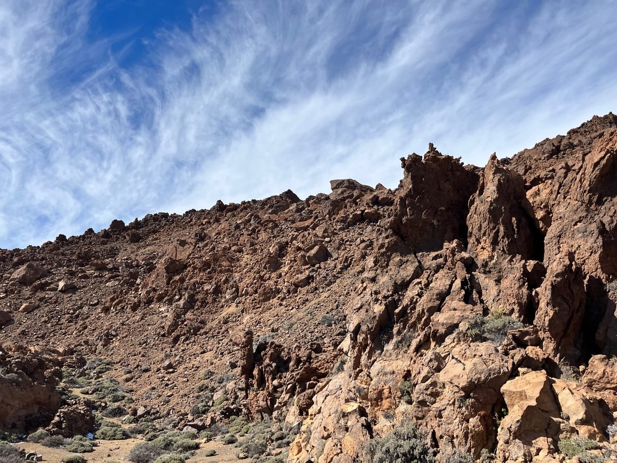 Steep reddish volcanic slope of jagged rocks under streaky white clouds