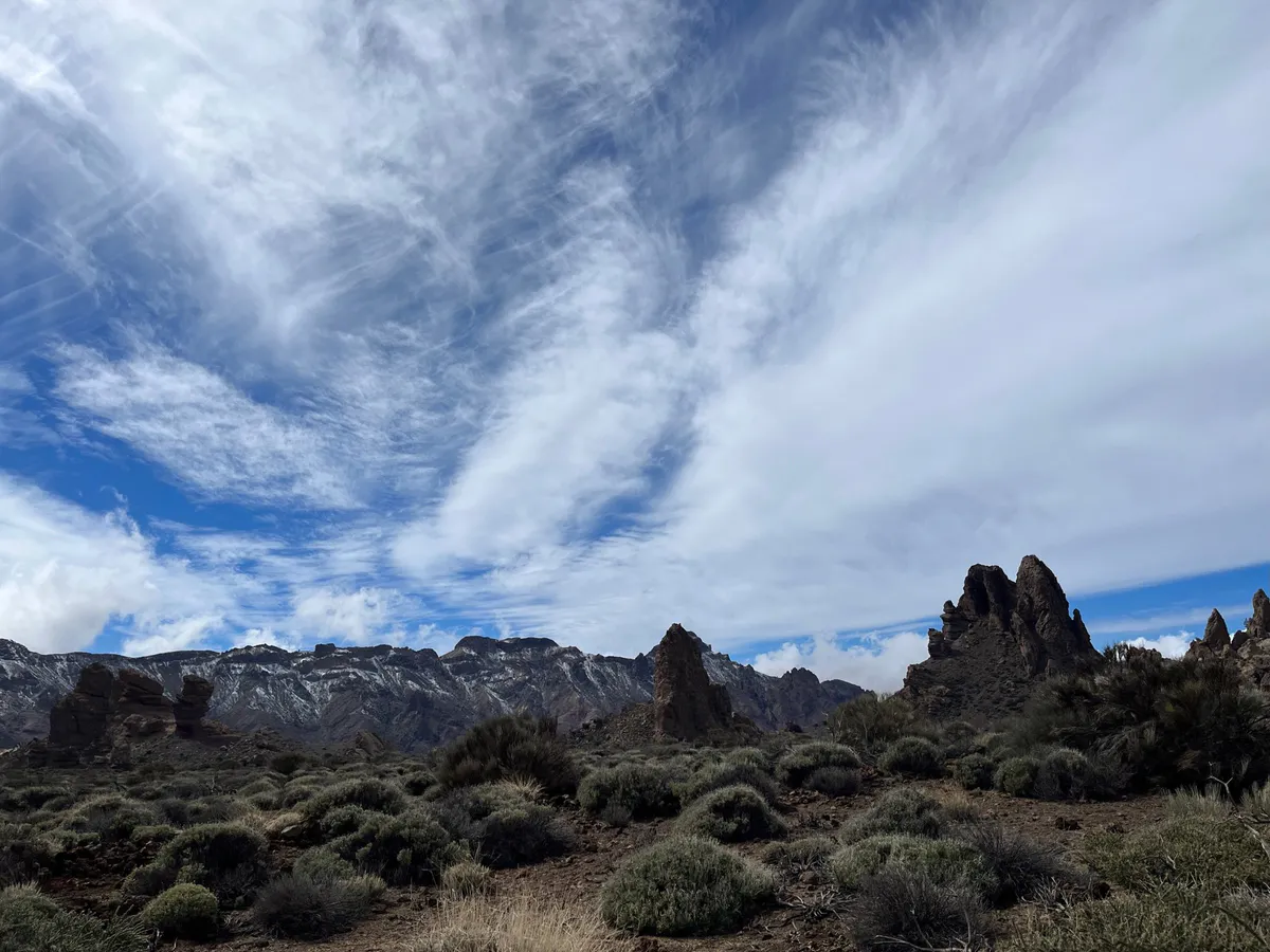 Rock pinnacles and snow-patched ridges above low shrubs under streaky clouds