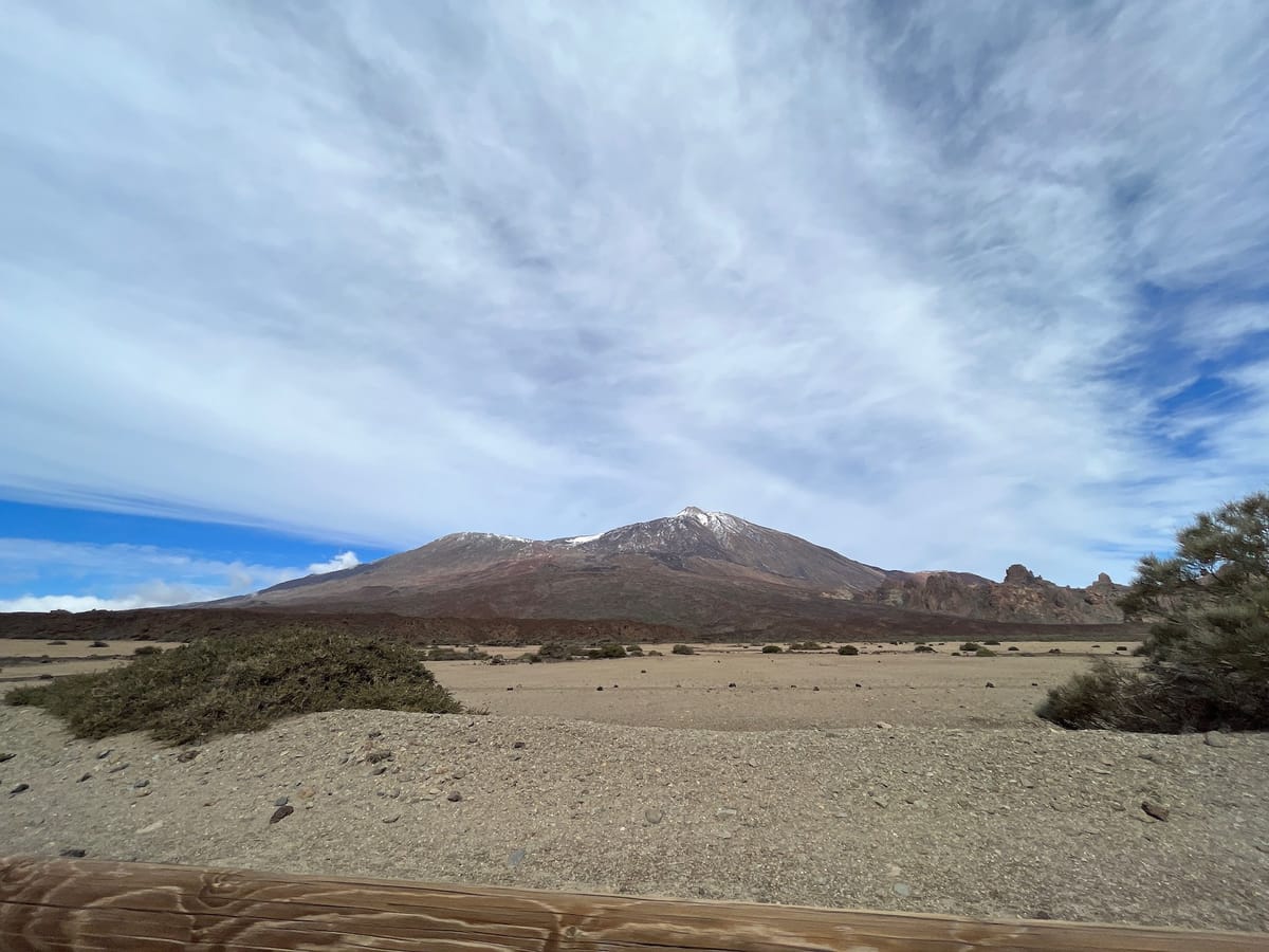 Snow-capped Teide across a sandy plain, seen over a wooden rail