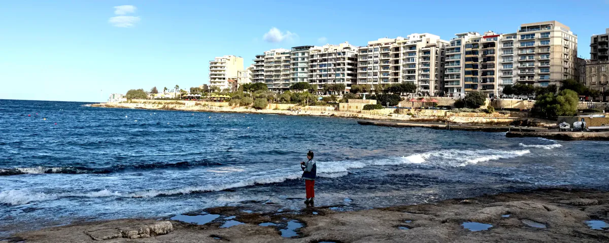 Person standing on a rocky Malta shoreline beside apartment blocks
