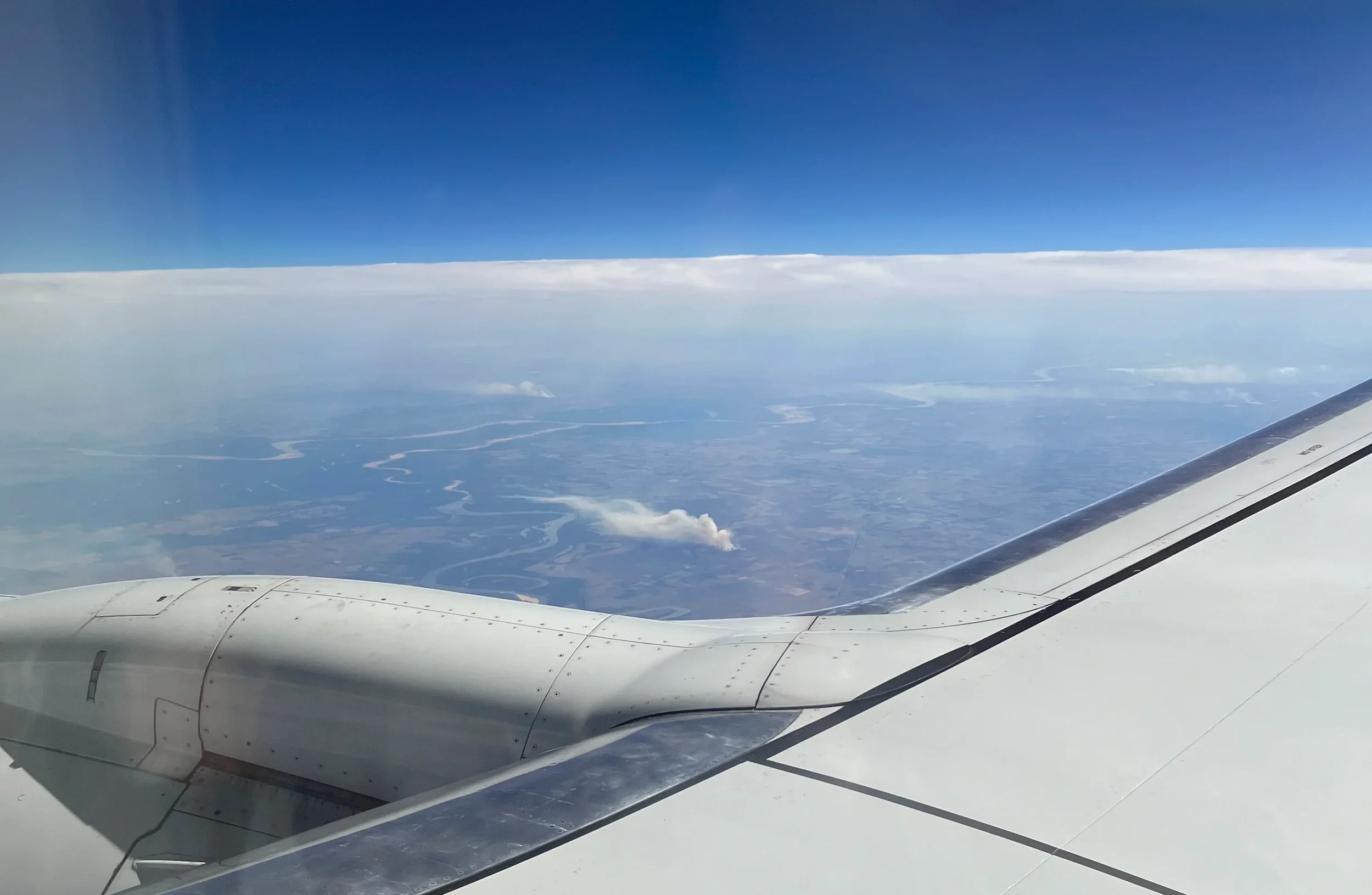 Airplane wing above a winding river, clouds, and hazy ground