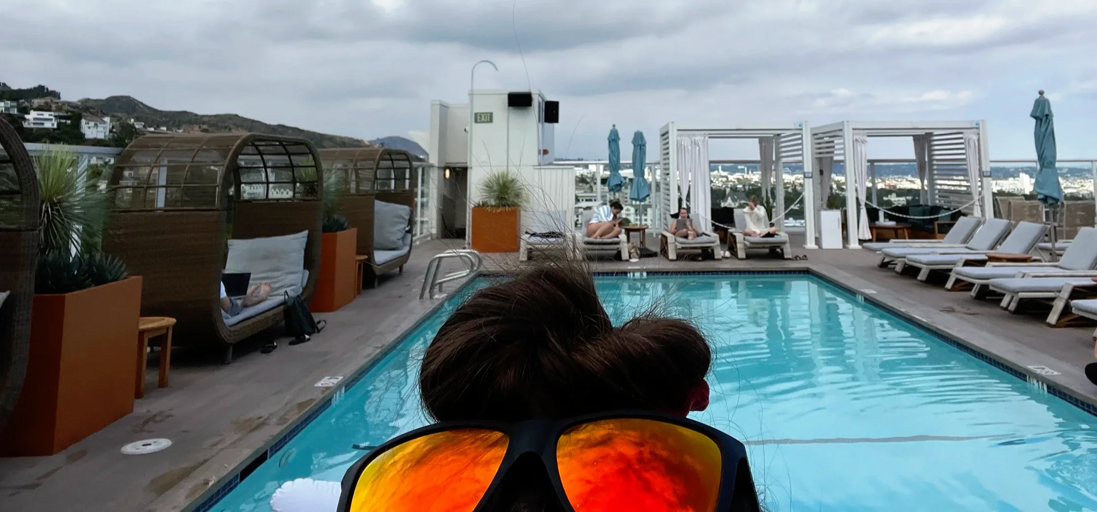 Person in orange sunglasses beside a rooftop pool and city view